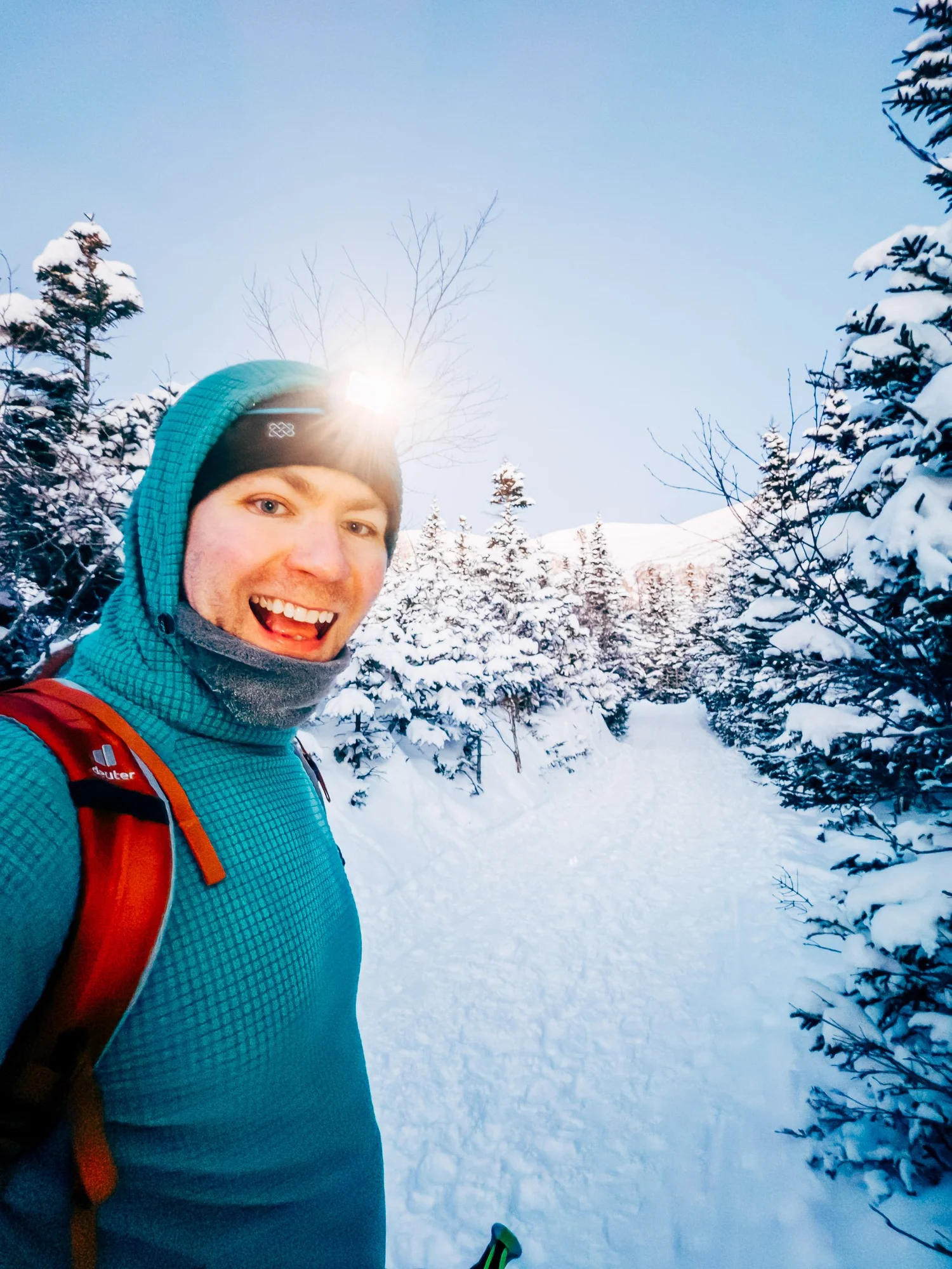 Smiling skier in a headlamp and teal hooded fleece on a snowy trail between frosted evergreens in early morning light
