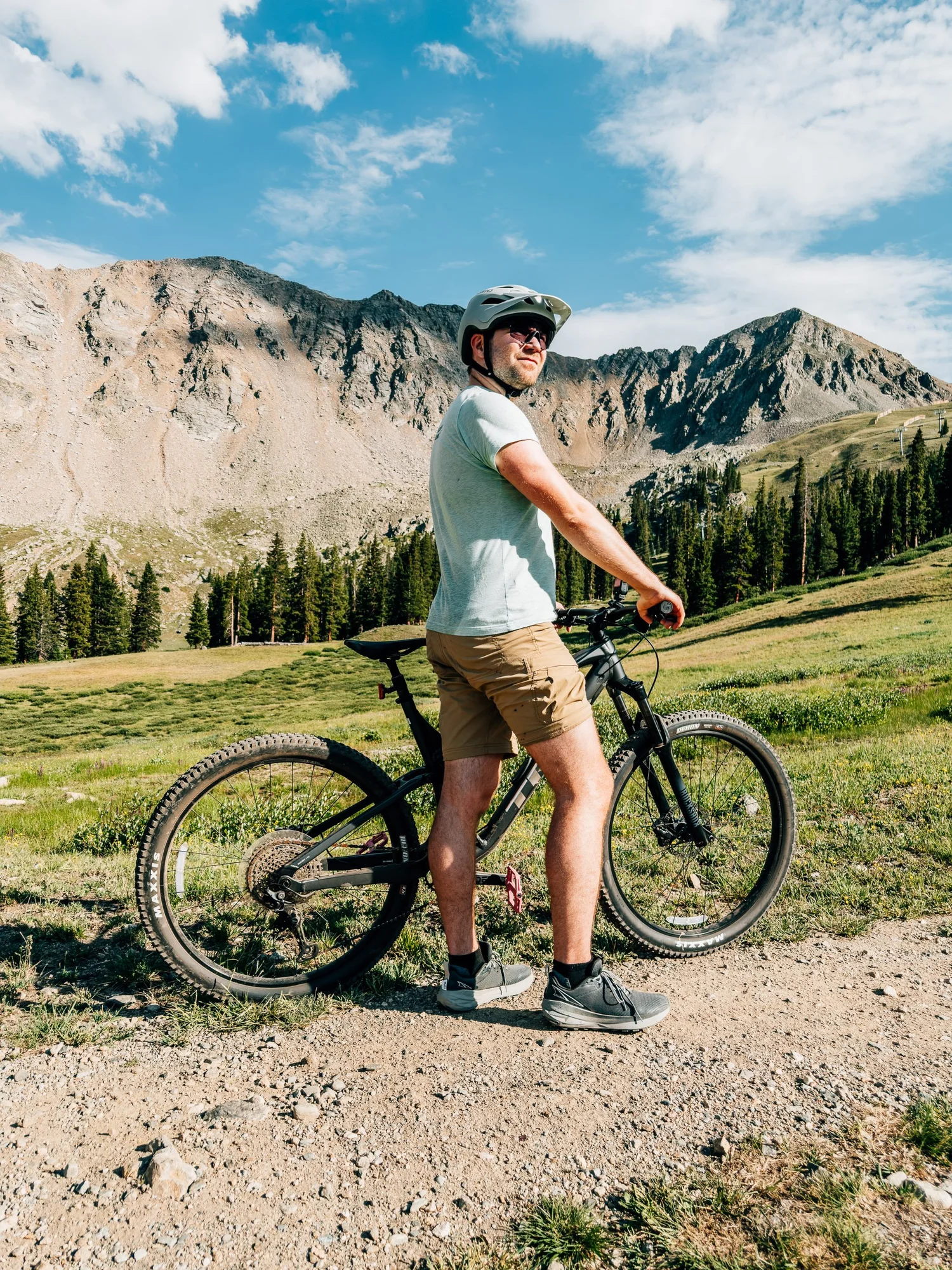 Mountain biker in a white helmet and sunglasses standing over his bike on a dirt road with rocky peaks and a green basin behind him