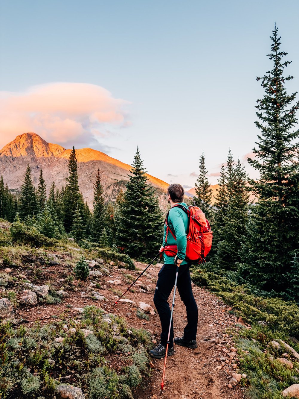 Hiker with an orange pack and trekking poles on a dirt trail looking toward a peak glowing orange at sunrise