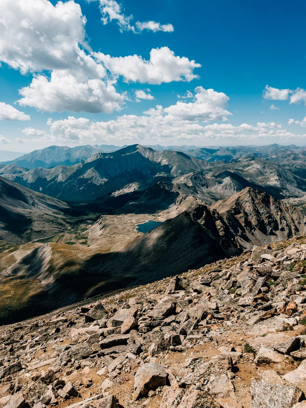 View from the rocky summit of Mount Harvard over a small alpine lake and layered mountain ranges under big clouds