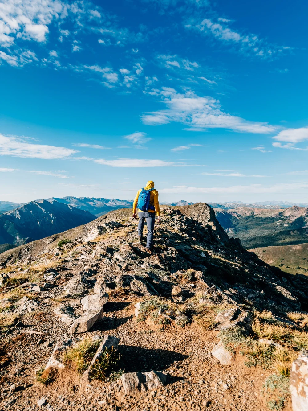 Hiker in a yellow hoodie and blue pack walking up a rocky ridge toward a wide mountain horizon under a deep blue sky