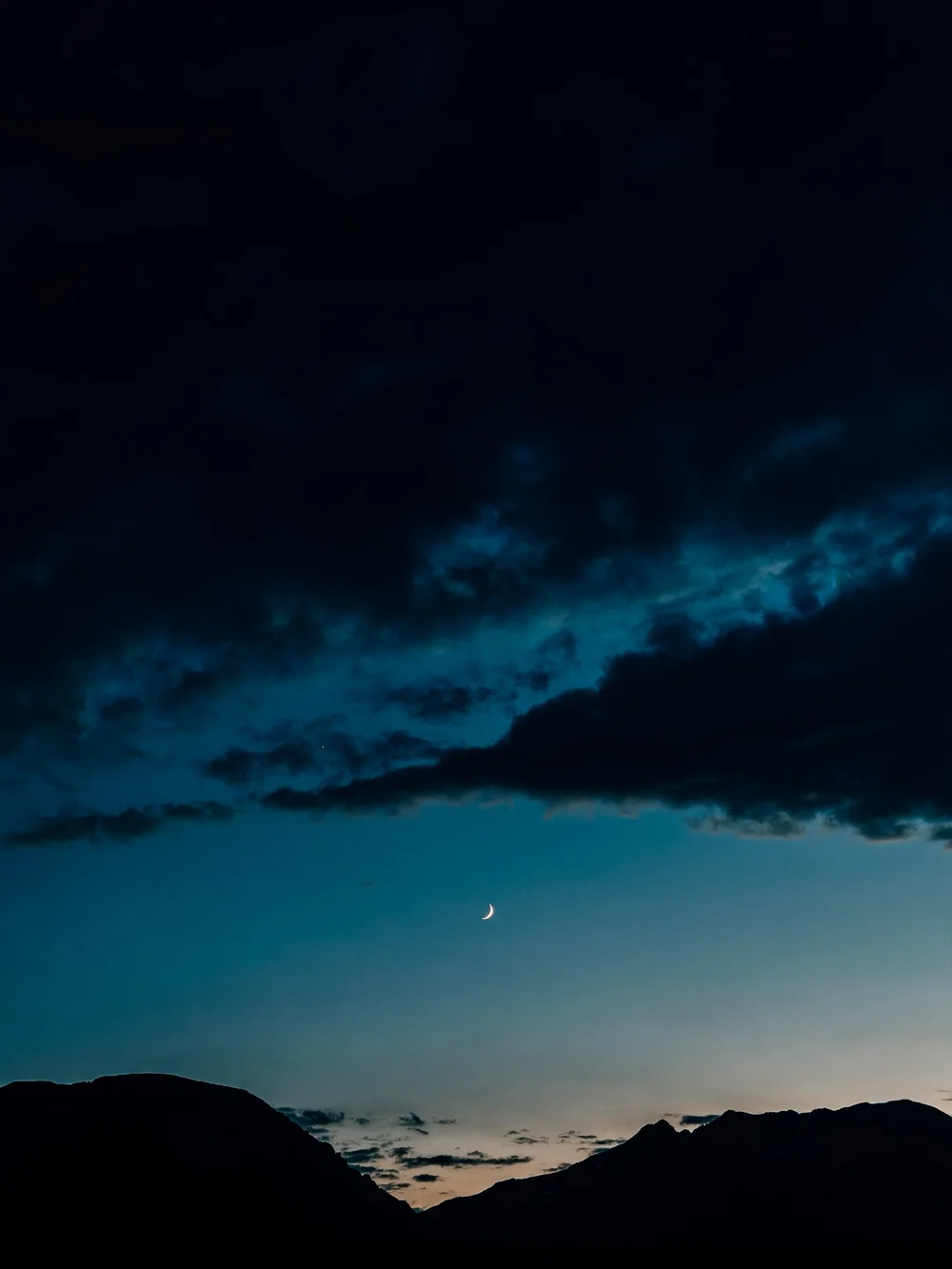 Thin crescent moon in a deep blue twilight sky above dark silhouettes of the Gore Range