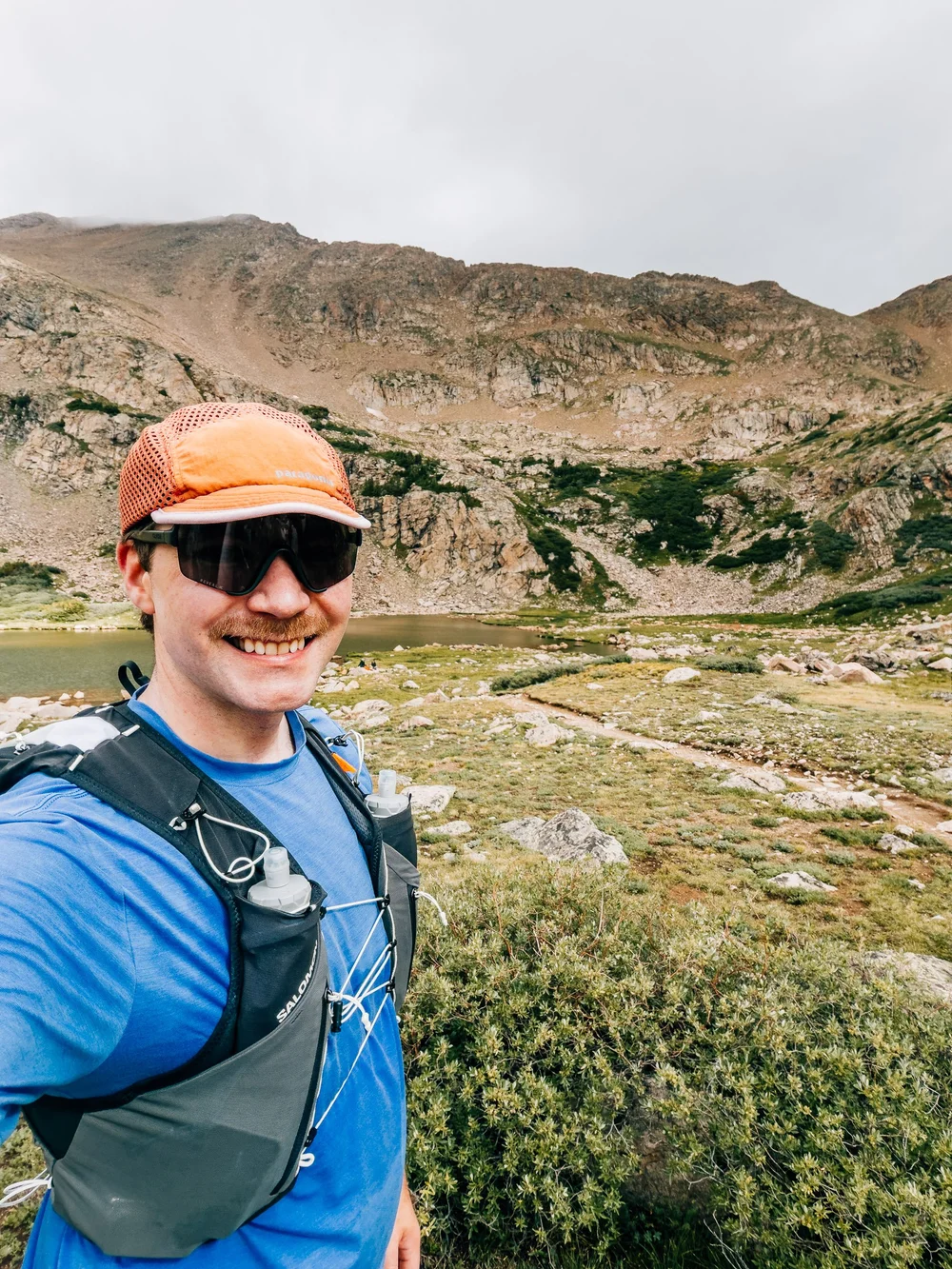Smiling trail runner in an orange cap and blue vest with an alpine lake and rocky basin behind him