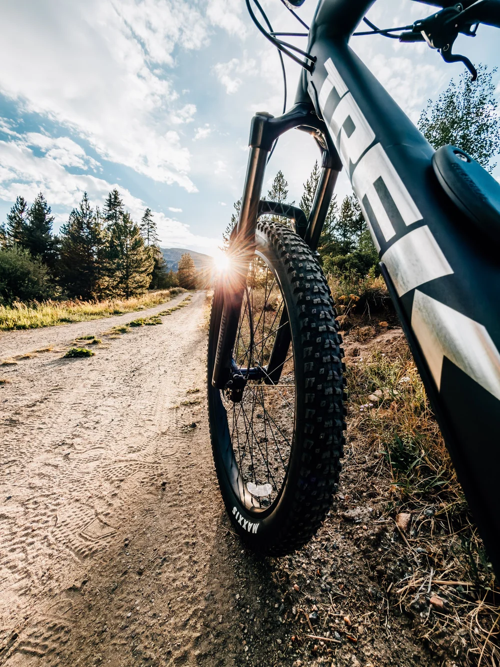 Low view of a mountain bike front wheel on a dirt road with the sun starburst through the trees at the end of the road