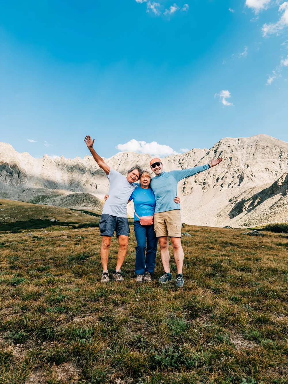 Three hikers with arms raised on an alpine meadow at Mayflower Gulch with rocky peaks and a bright blue sky behind them