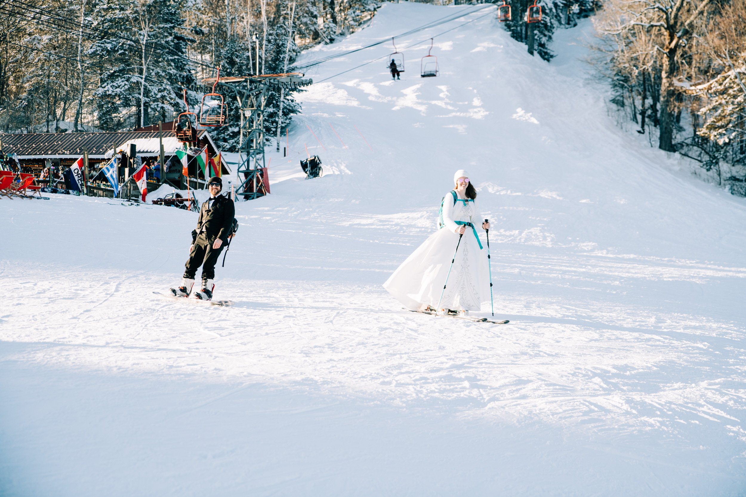 Bride in a wedding dress and groom skiing down the slope at Black Mountain in New Hampshire past the chairlift