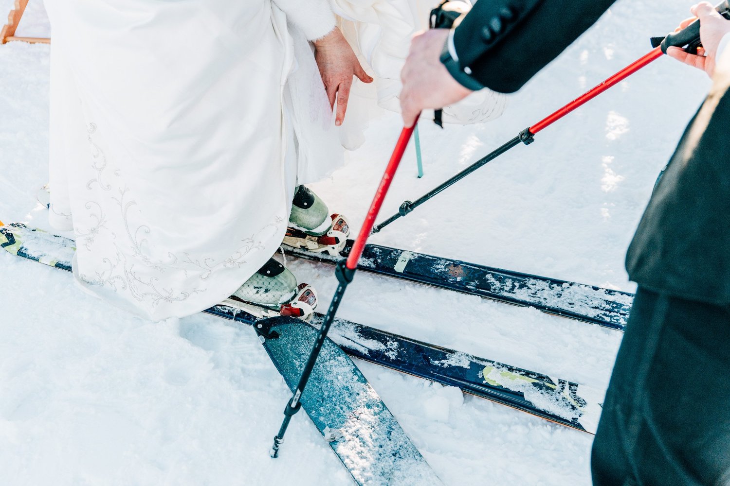 Bride in a wedding dress and groom clipping into skis with ski poles during a New Hampshire ski elopement