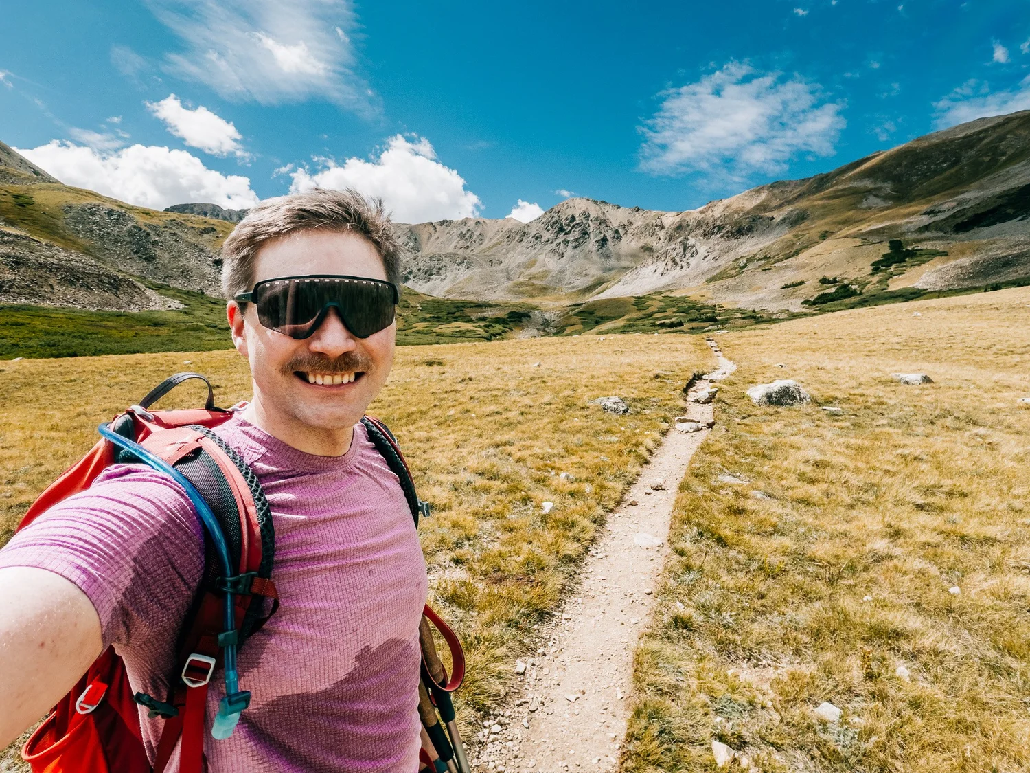 Anton grinning in sunglasses on the trail through the golden upper basin of Missouri Gulch, rocky peaks behind