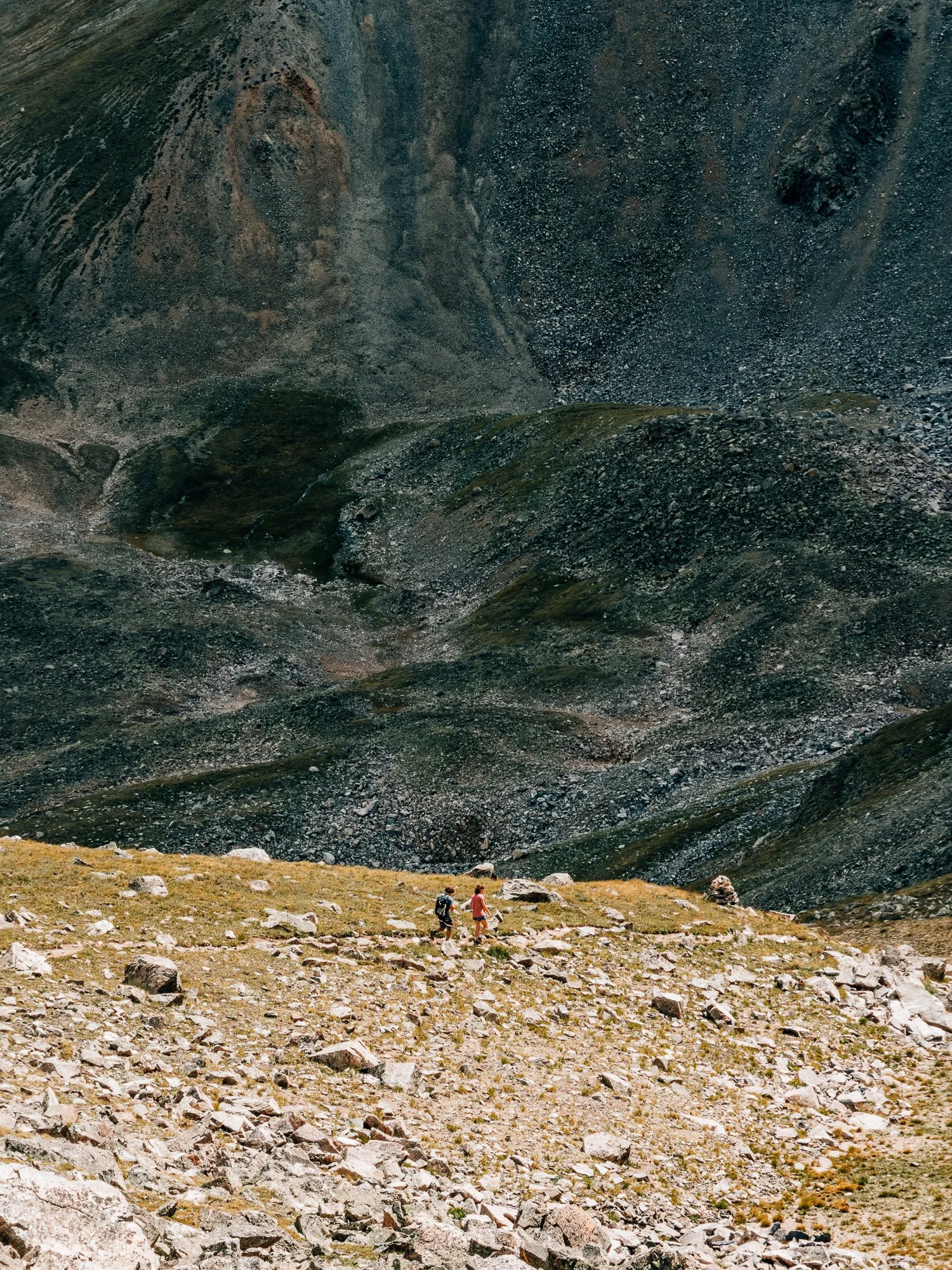 Two small hikers crossing a tundra slope below a huge dark scree face on Missouri Mountain