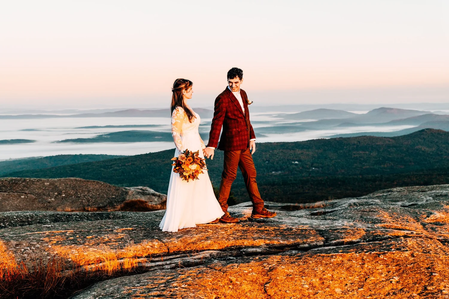 Couple walking a granite summit ledge at sunrise above a valley filled with fog in New Hampshire