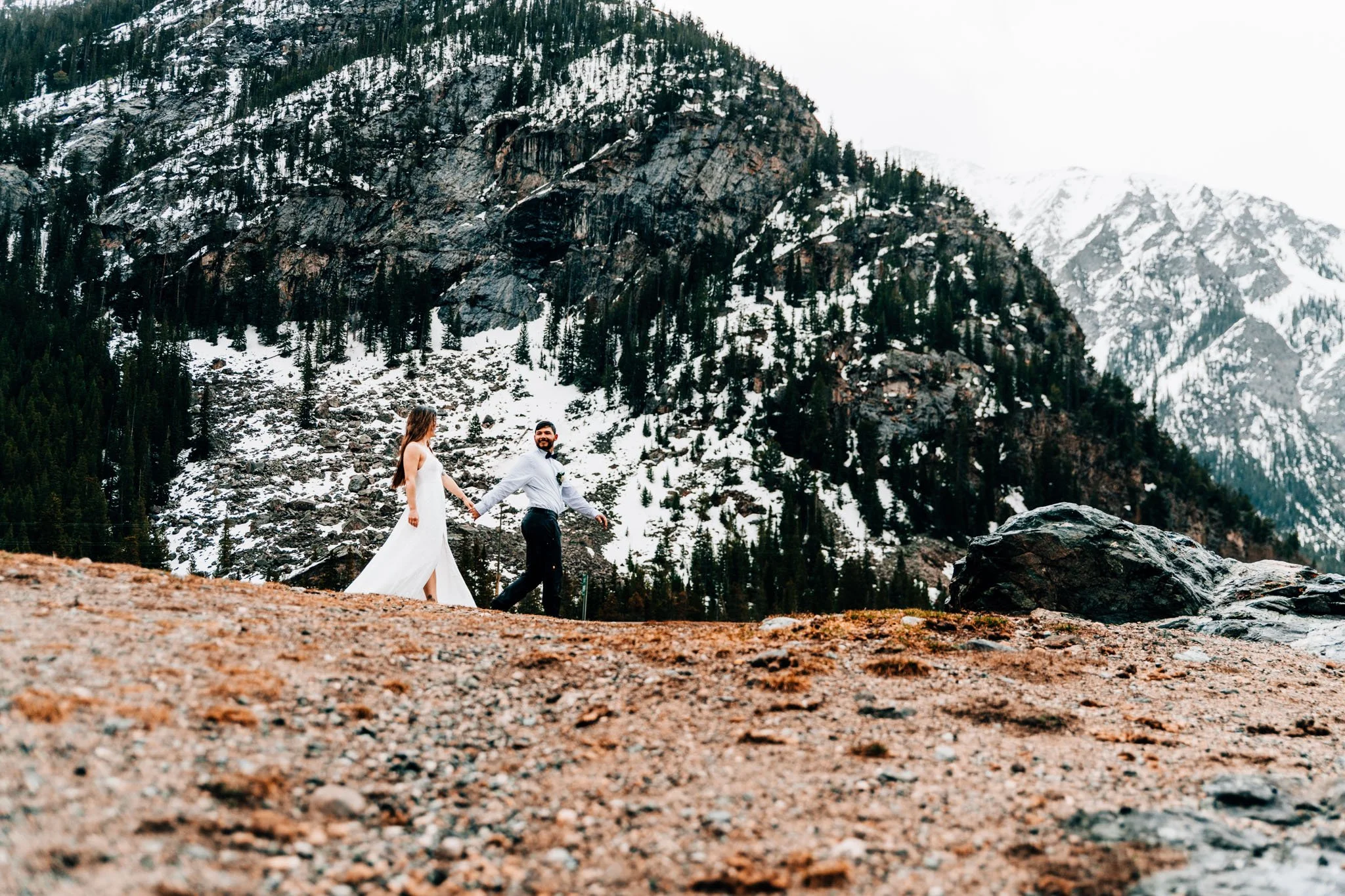 Couple crossing an open rocky shoreline with snowy Colorado peaks rising behind them