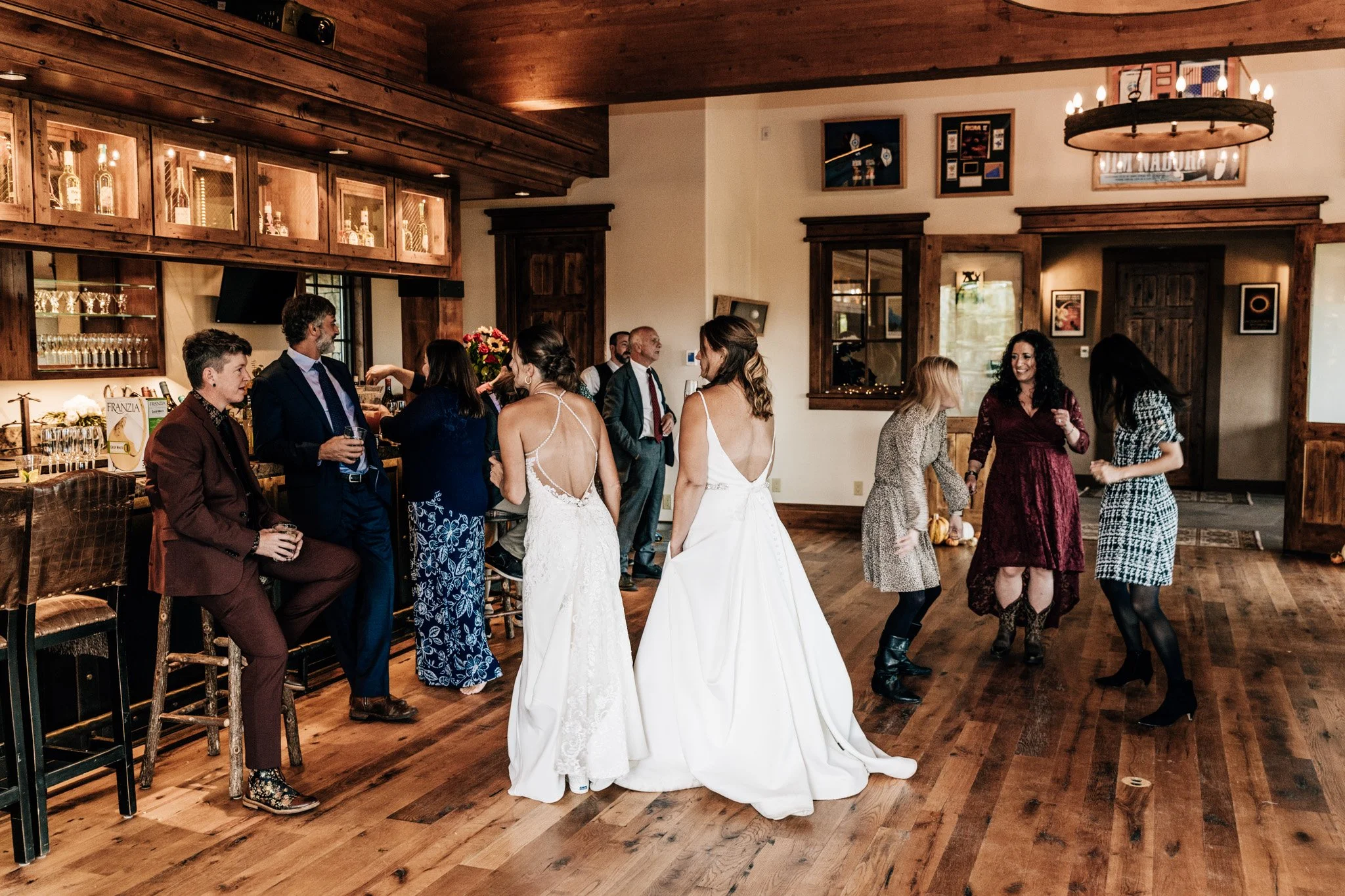 Two brides in white gowns mingling with their guests at an intimate wedding reception inside a timber-framed lodge