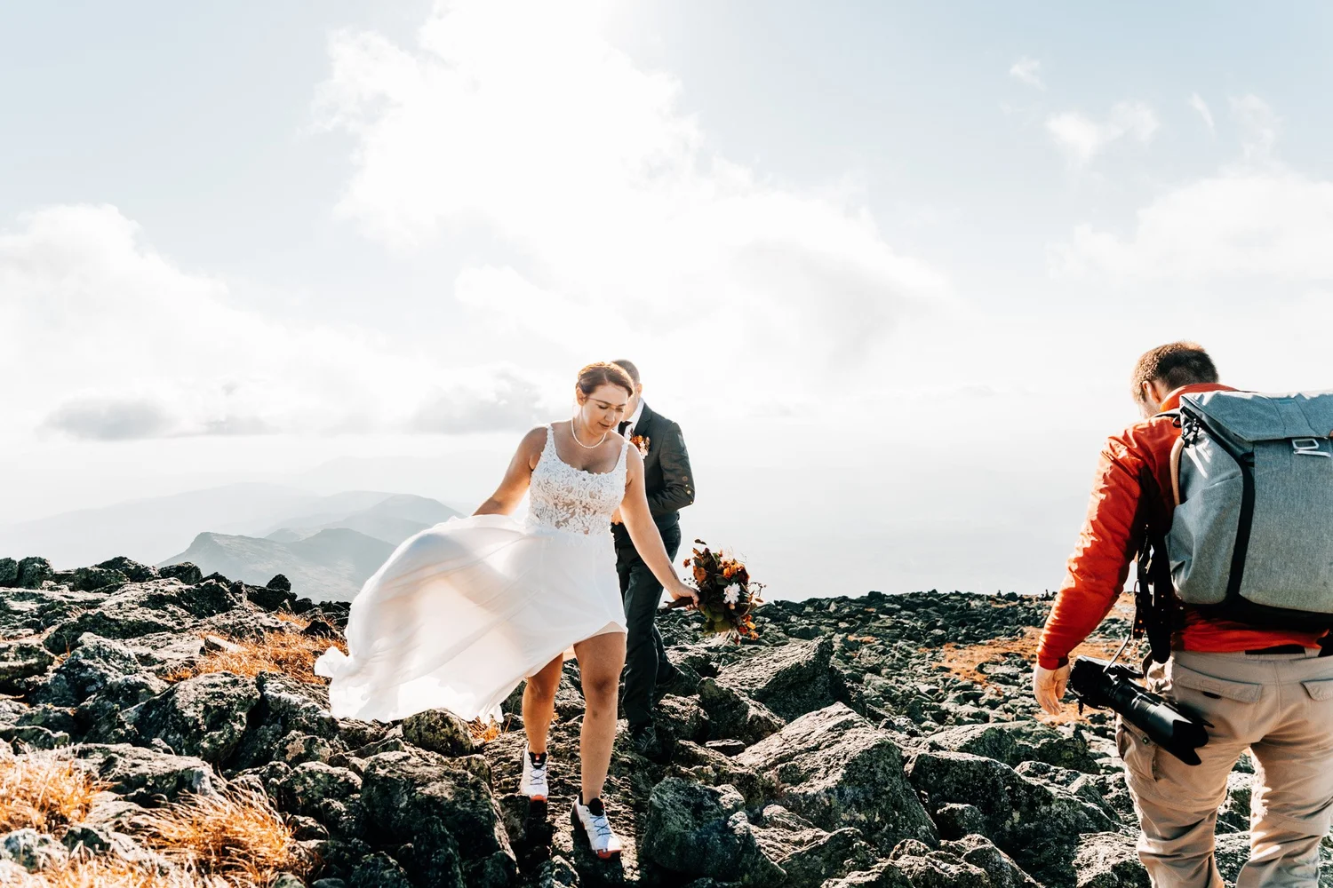 Danielle and Tom picking their way across the rocky summit of Mount Washington on their October wedding day, photographer following behind