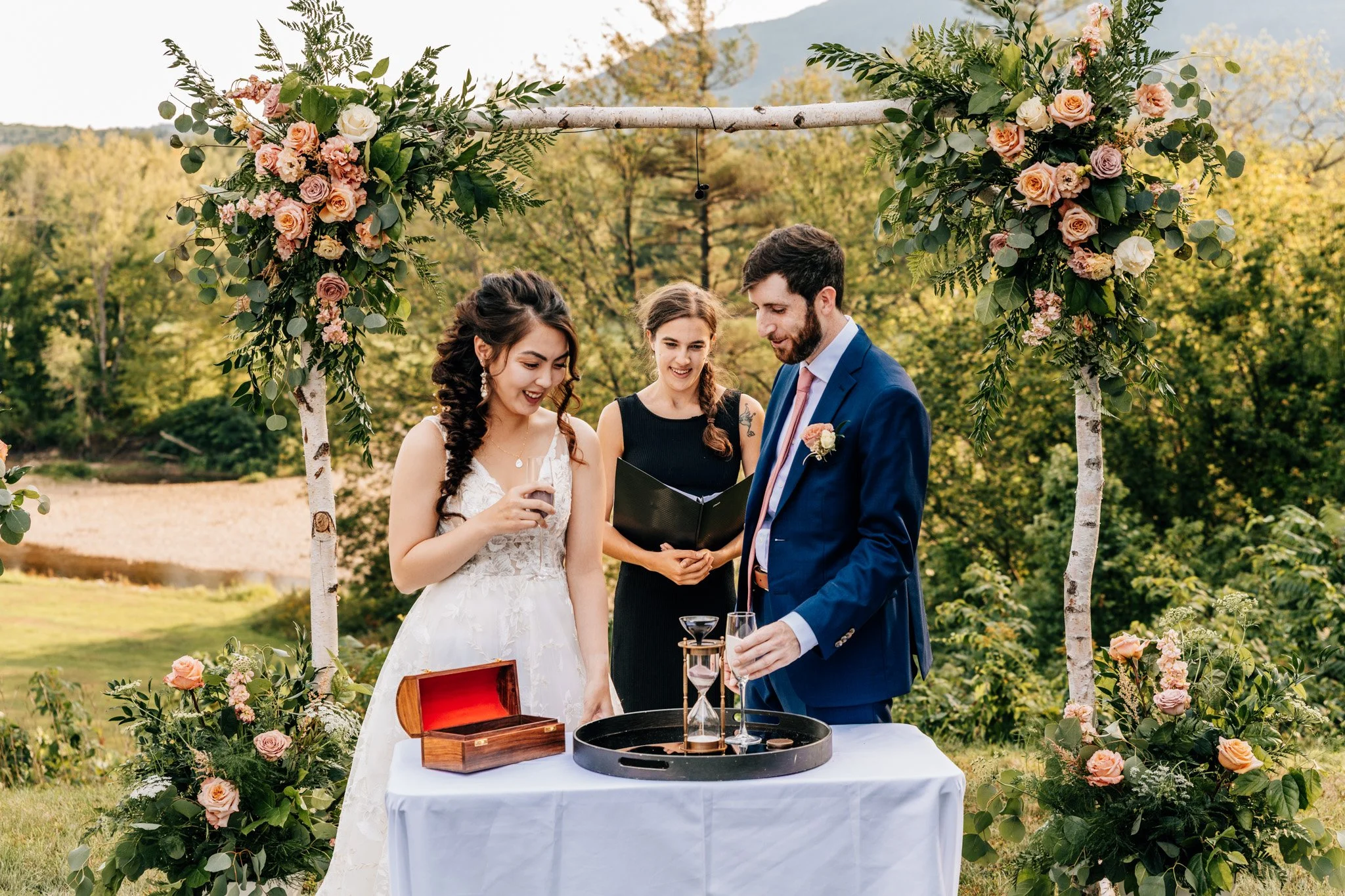 Couple and officiant at a small outdoor wedding ceremony beneath a birch-branch arch of peach roses in North Conway, New Hampshire