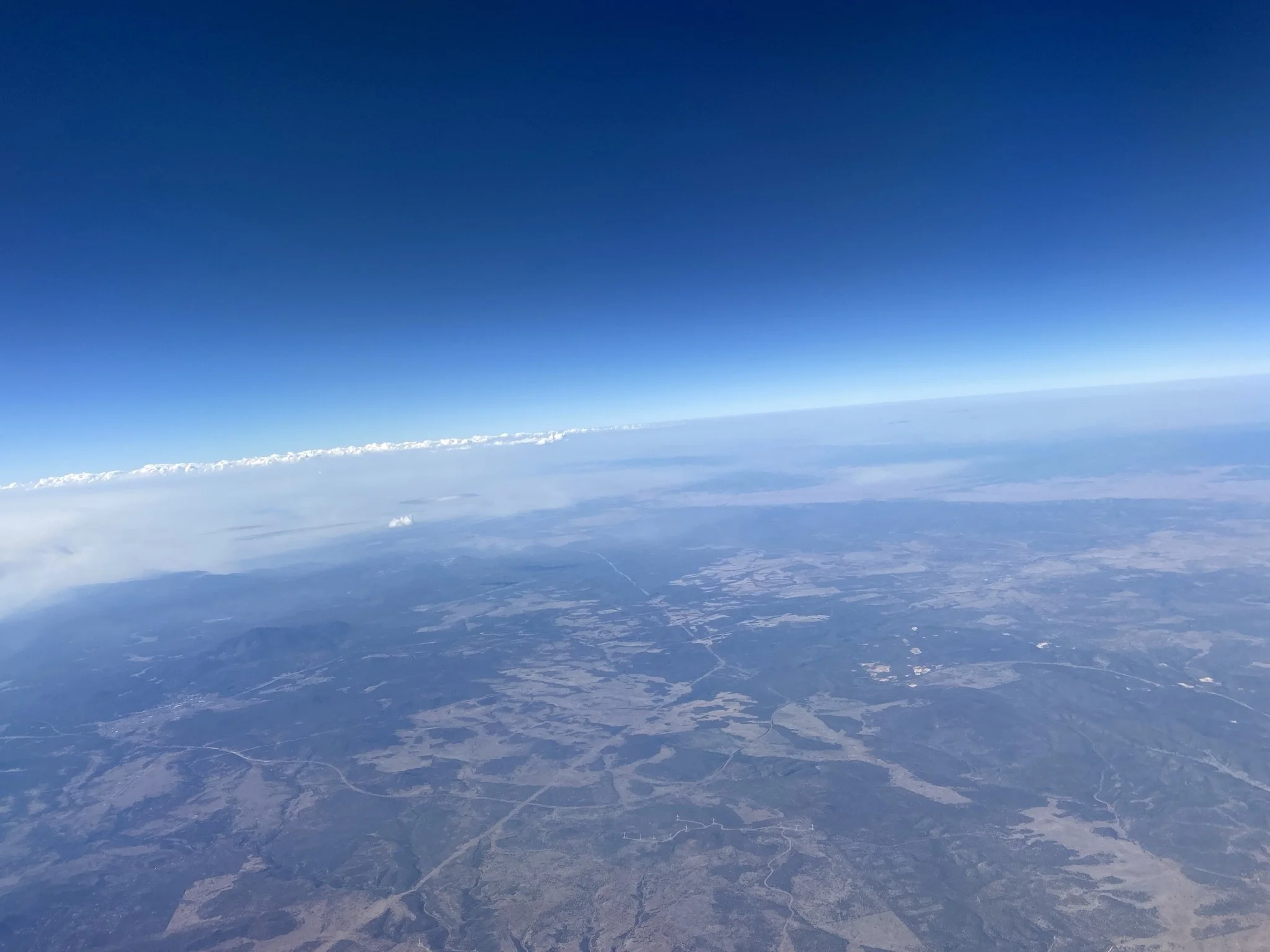 Aerial view of Earth's surface showing land with patches, water bodies, and distant mountains under a clear blue sky with some clouds near the horizon.