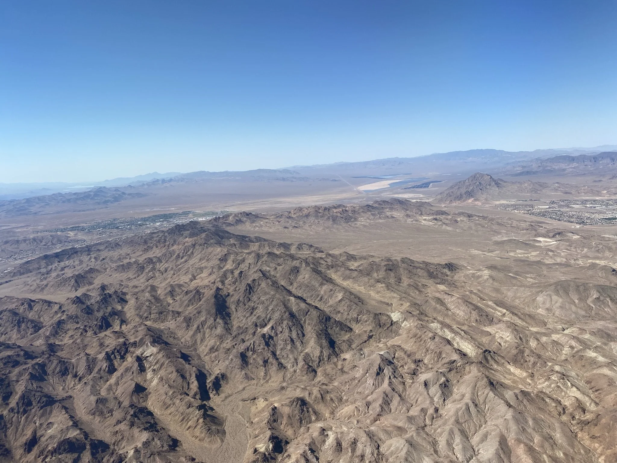 Aerial view of a desert landscape with rugged mountains, flat plains, and distant mountains under clear blue sky.