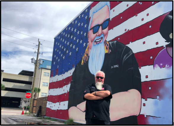 Ray “Dr. BBQ” Lampe standing in front of the Dr. BBQ mural by Cam Parker (aka Painkiller Cam)