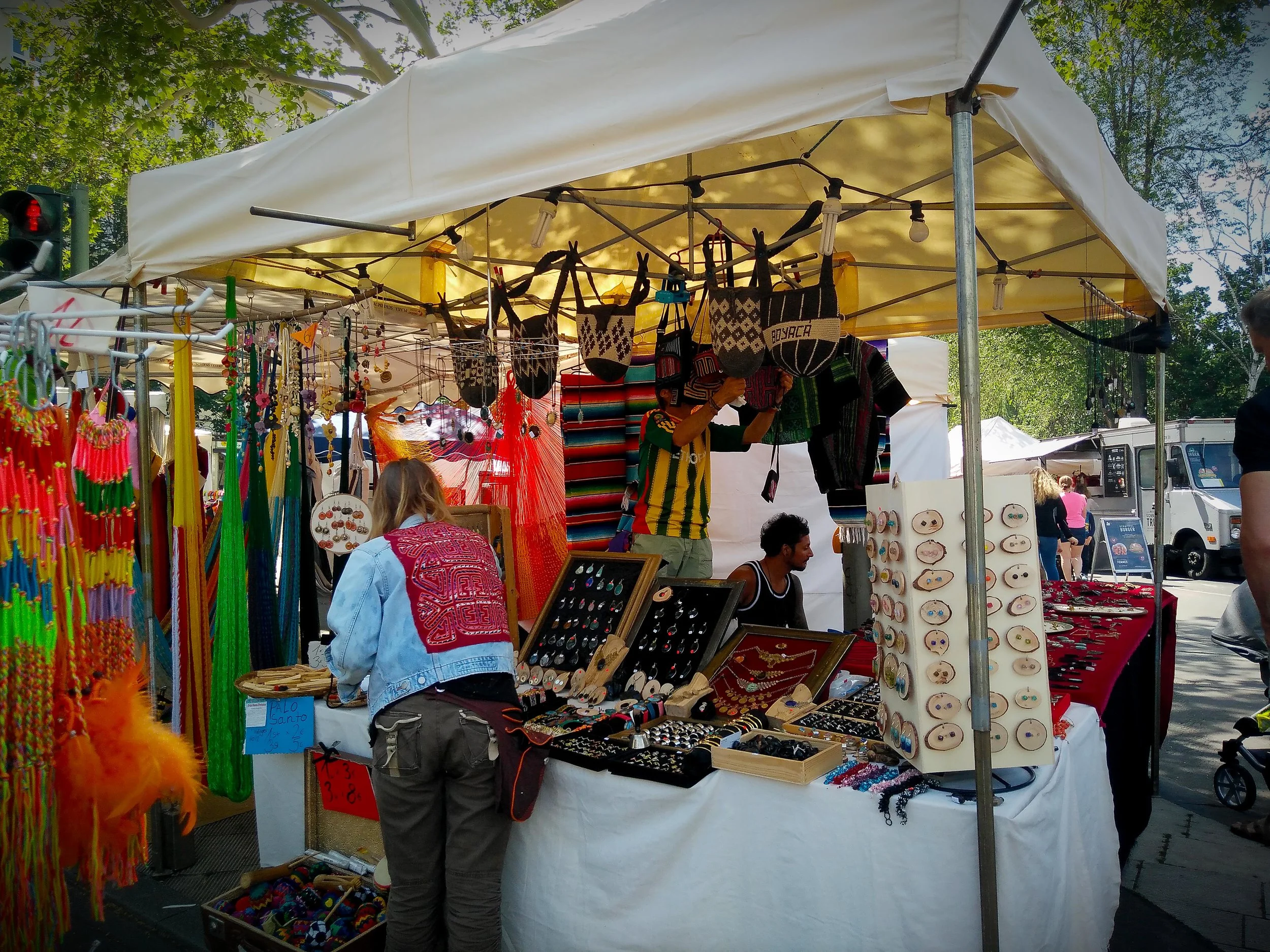 Carnival of Cultures, Berlin, Germany 2019. Vendor selling hand braided bracelets, trinkets, necklaces, and other accessories. The jean sweater has a mola textile attached on the back.