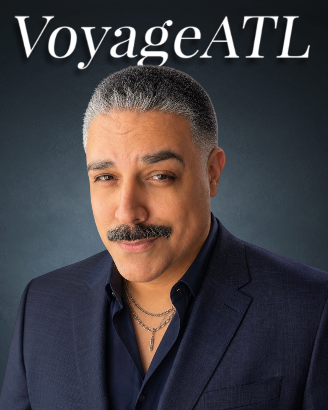 A middle-aged man with short, gray hair, a mustache, wearing a dark suit jacket, a dark shirt, and layered silver necklaces, looking at the camera with a confident expression. The background is dark and textured, with the text 'VoyageATL' above his head.
