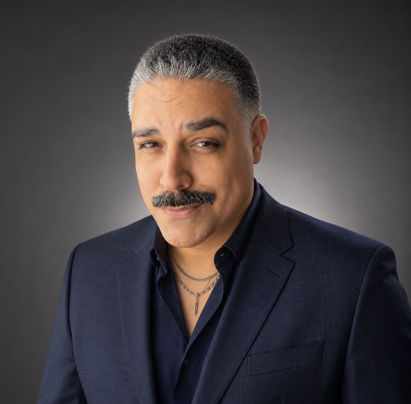 Headshot of a man with gray hair, a mustache, and light complexion, wearing a dark blazer, navy shirt, and layered necklaces, smiling slightly against a gray gradient background.