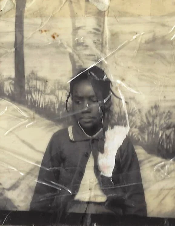 A young girl with braided hair and a serious expression, wearing a dark jacket over a collared shirt, standing in front of a painted backdrop of trees and grass. The photo is covered with cracks or scratches.