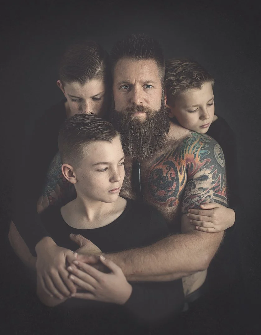 A man with a beard and tattoos is surrounded by three children, all embracing him, against a dark background. This is a photo by one of the top photographers in hawke's bay, Eva Bradley