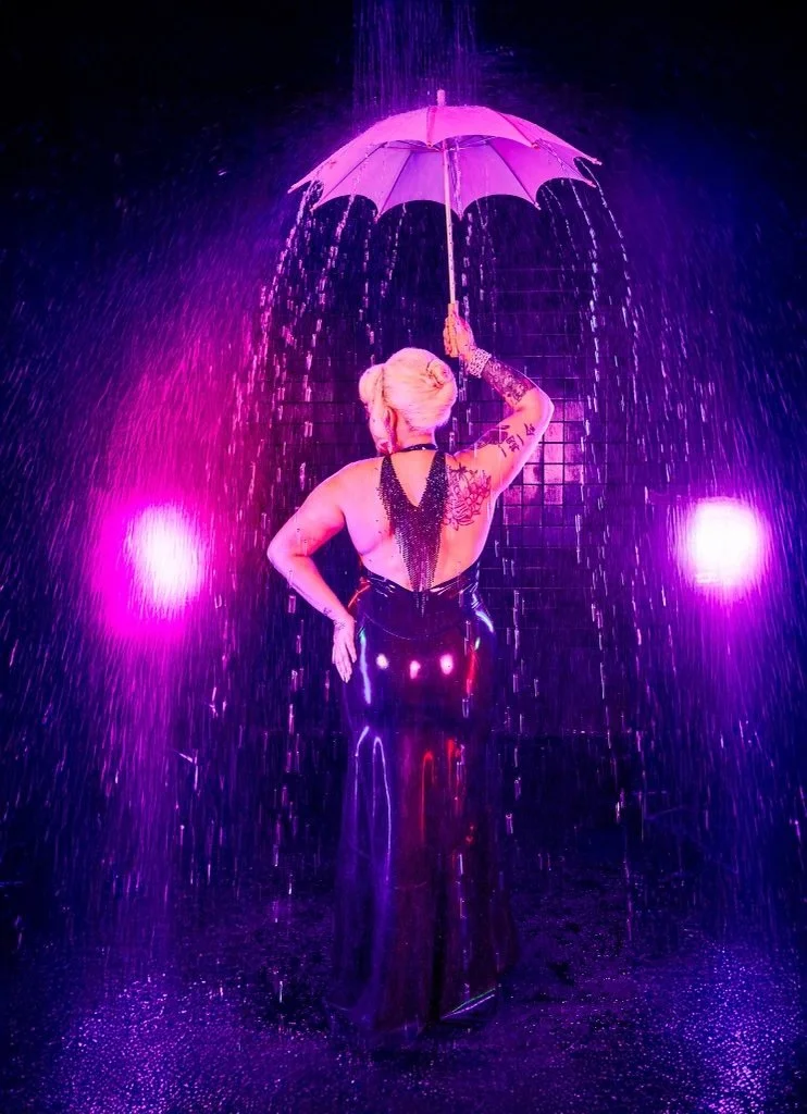 Person in a black gown under an umbrella in dramatic purple rain lighting