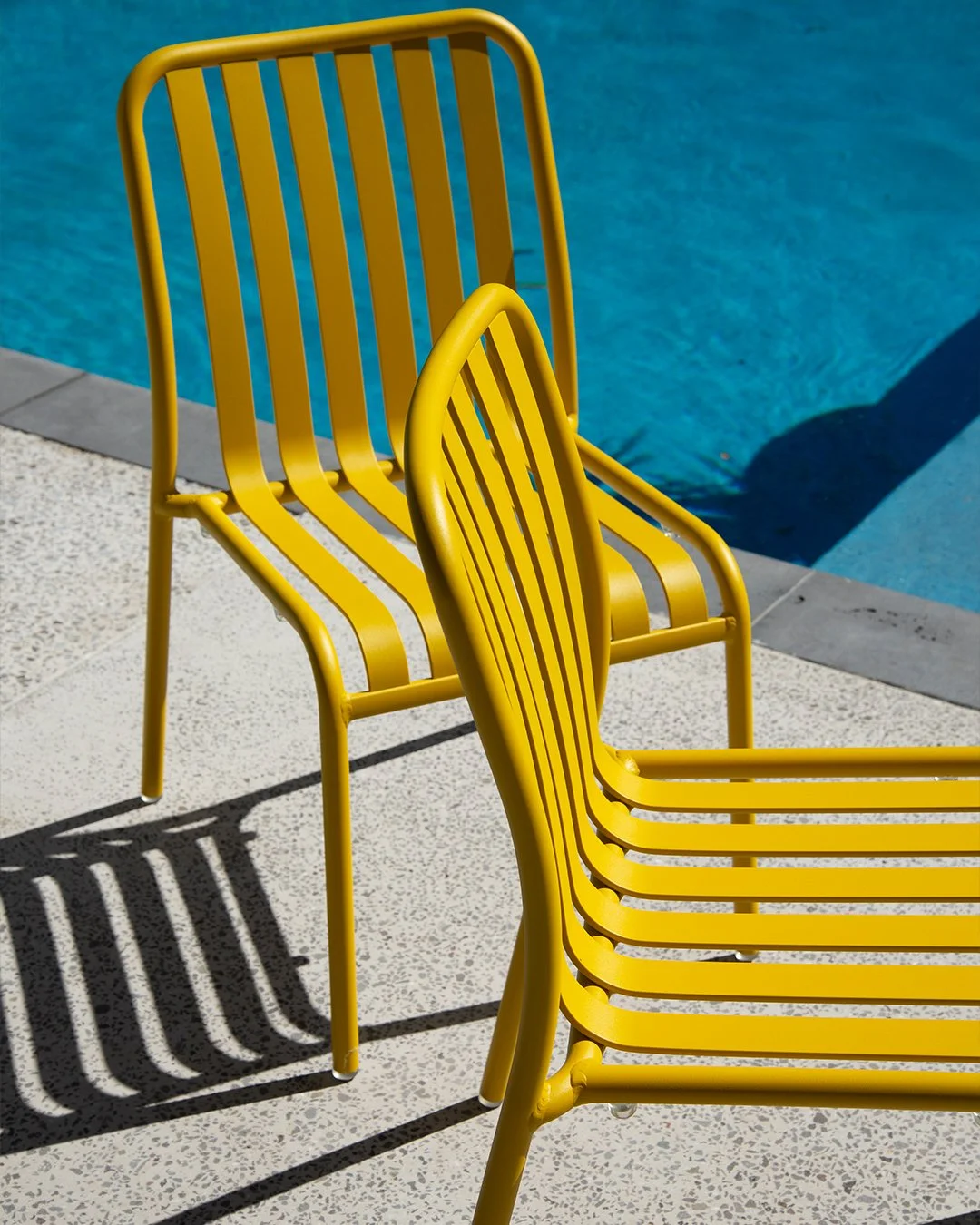 Yellow metal poolside chairs casting shadows on concrete near a blue swimming pool.