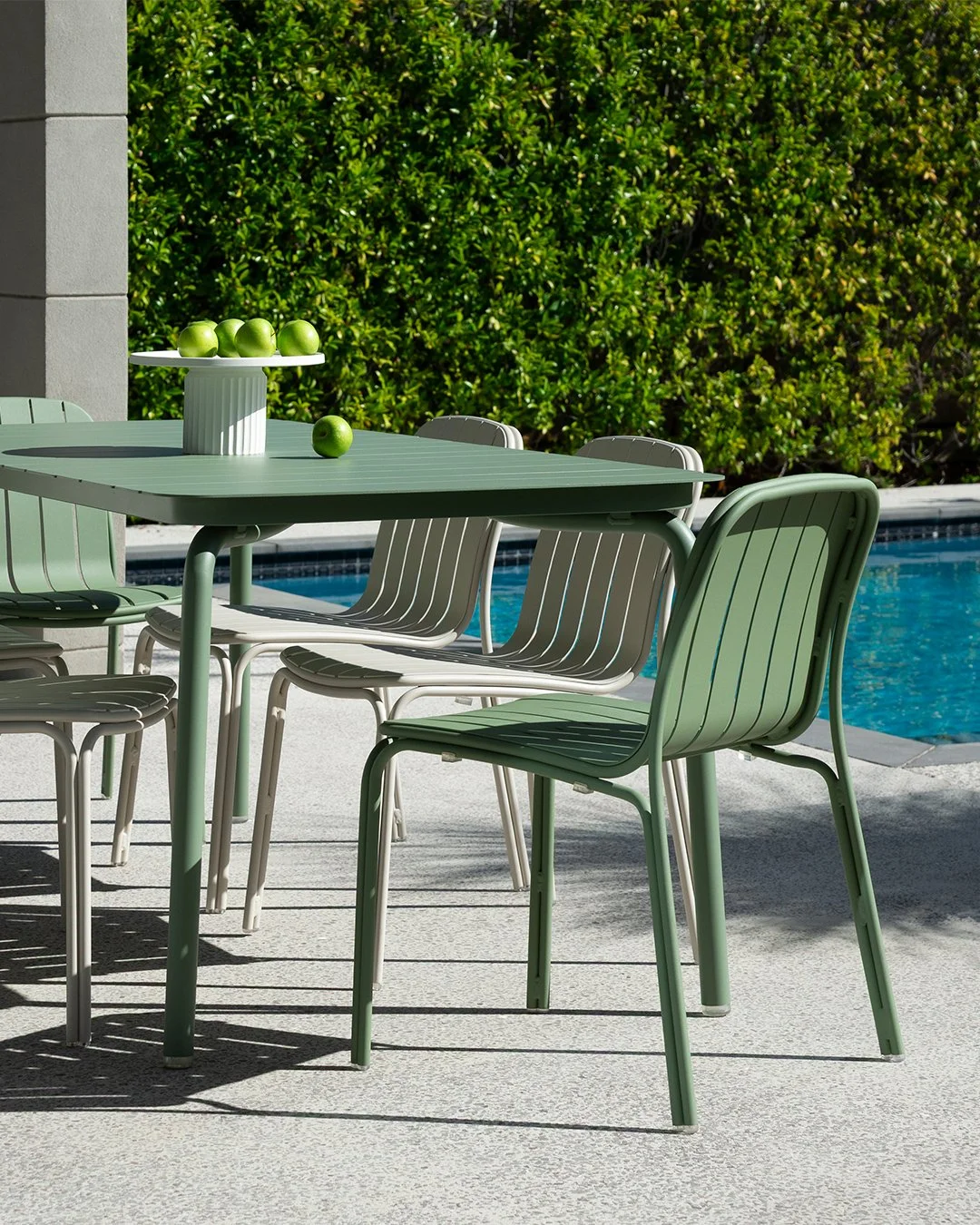 Green patio table with matching chairs, a white vase with green apples, near a swimming pool with green hedge in background.