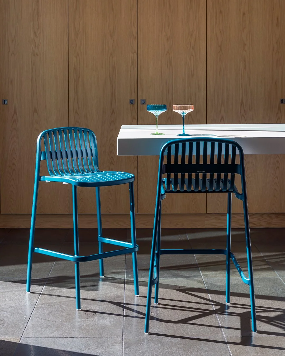 Two blue metal chairs and a white table with two colorful glasses on it, set against a wooden cabinet background.