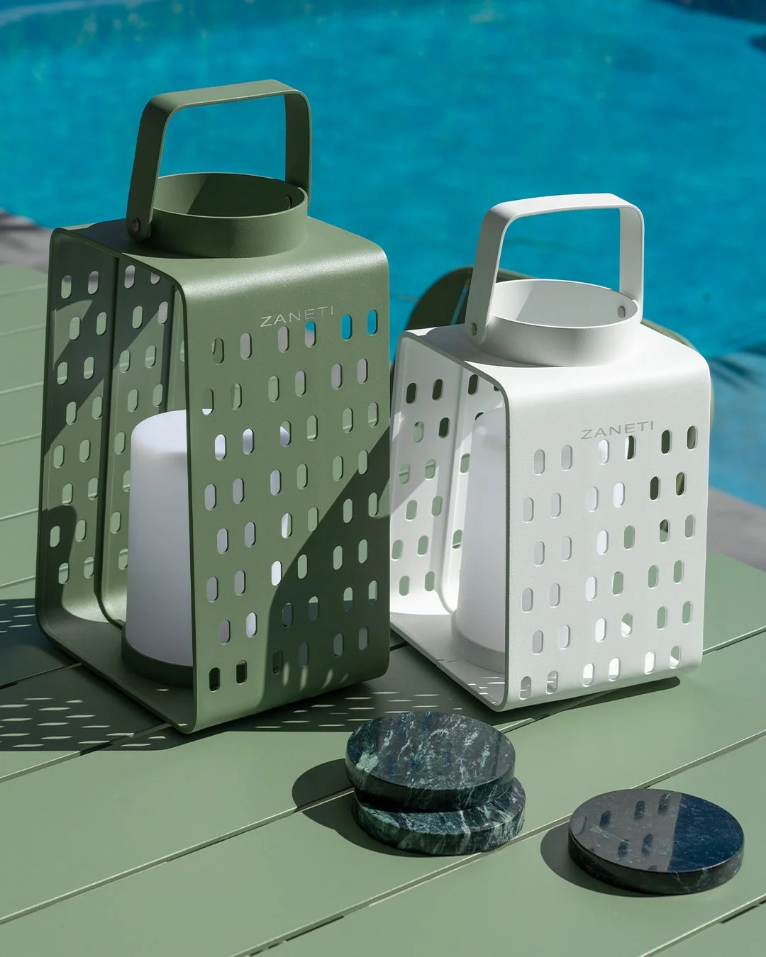 Two modern lanterns, one green and one white, with the brand name 'ZANETI' written on them, are placed on a green outdoor table near a swimming pool. There are also two black and green marble coasters nearby.