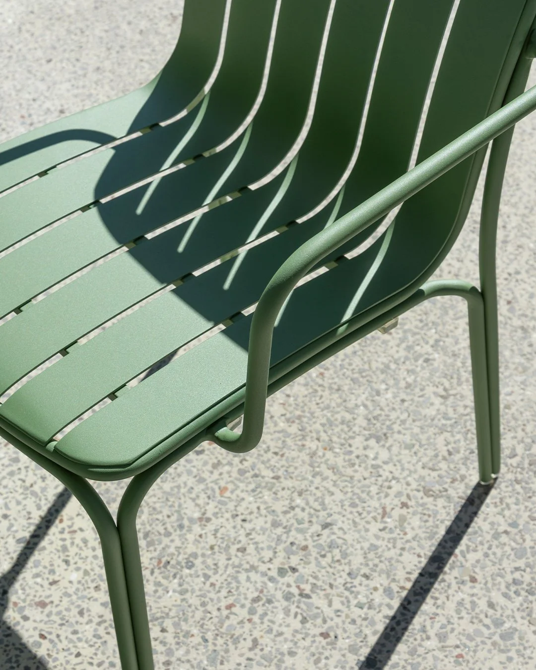 A mint green outdoor metal chair with slatted design, casting shadows on a speckled concrete ground.