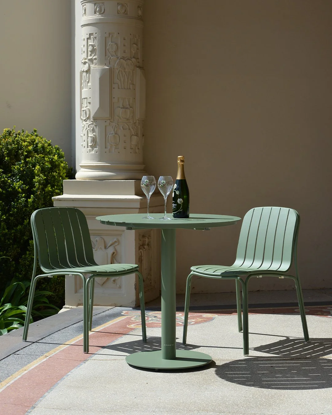 Outdoor patio scene with a green table, two matching chairs, a bottle of champagne, and two empty champagne glasses, with decorative architecture and a bush in the background.