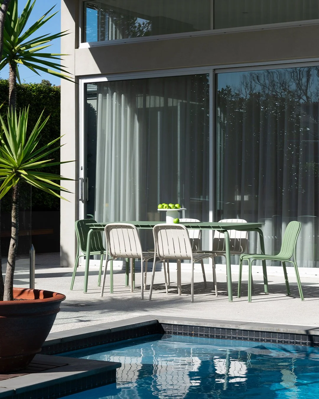 Outdoor patio with a green and beige dining table set near a swimming pool, with a modern house featuring large glass sliding doors and white curtains in the background. Potted palm trees are also visible.