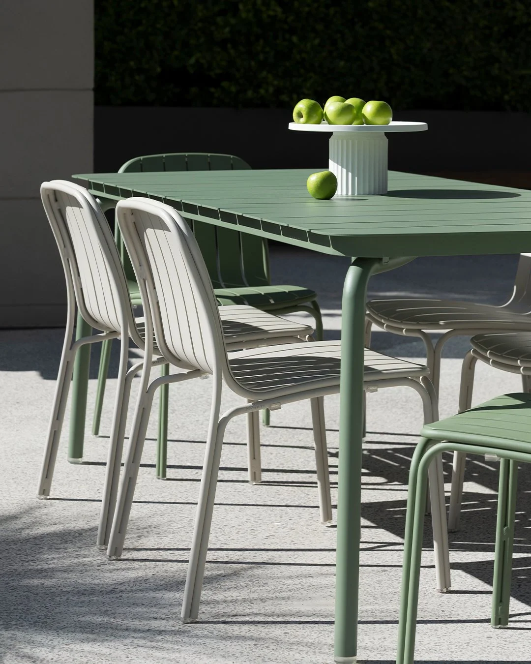 Outdoor patio with a light green table surrounded by white and green chairs, and a white cake stand with green apples on top, one apple on the table.