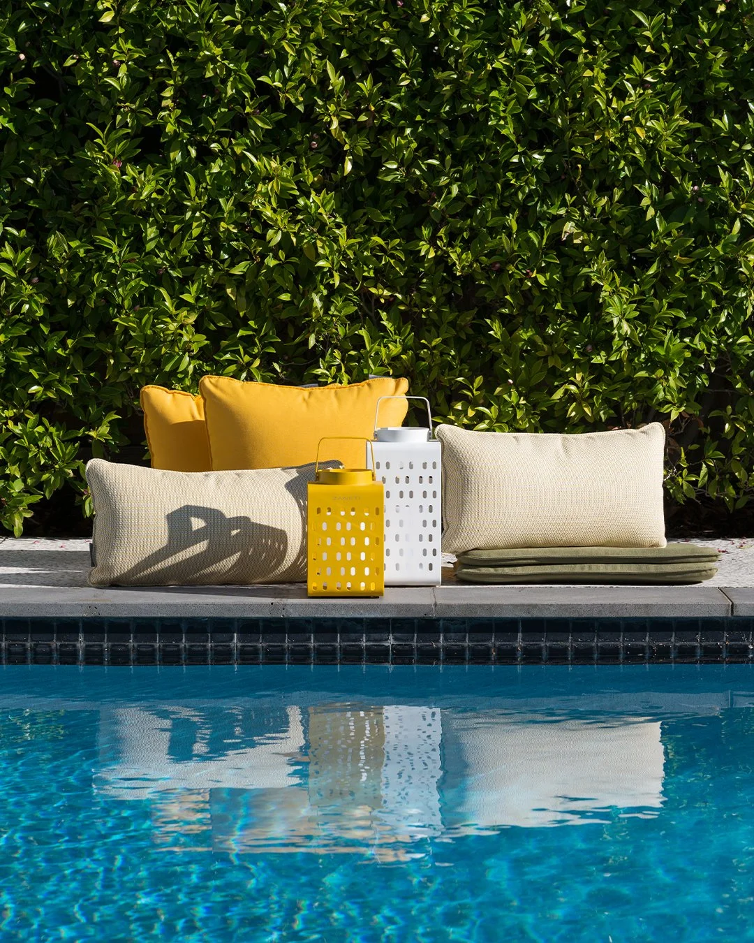 Colorful pillows and lanterns near a swimming pool surrounded by lush green foliage.
