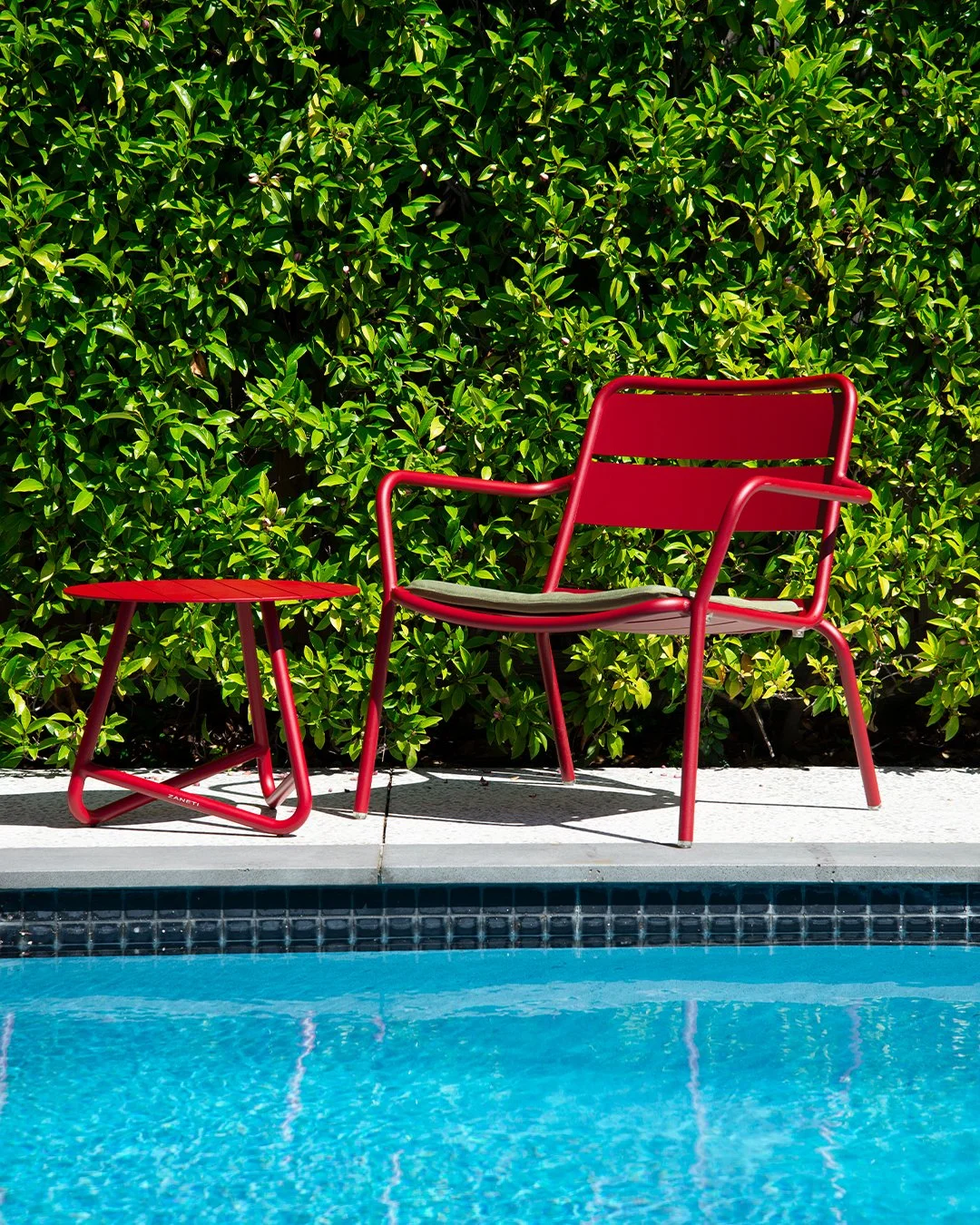 A red chair and a small red table beside a swimming pool with blue water, set against a backdrop of green shrubbery.