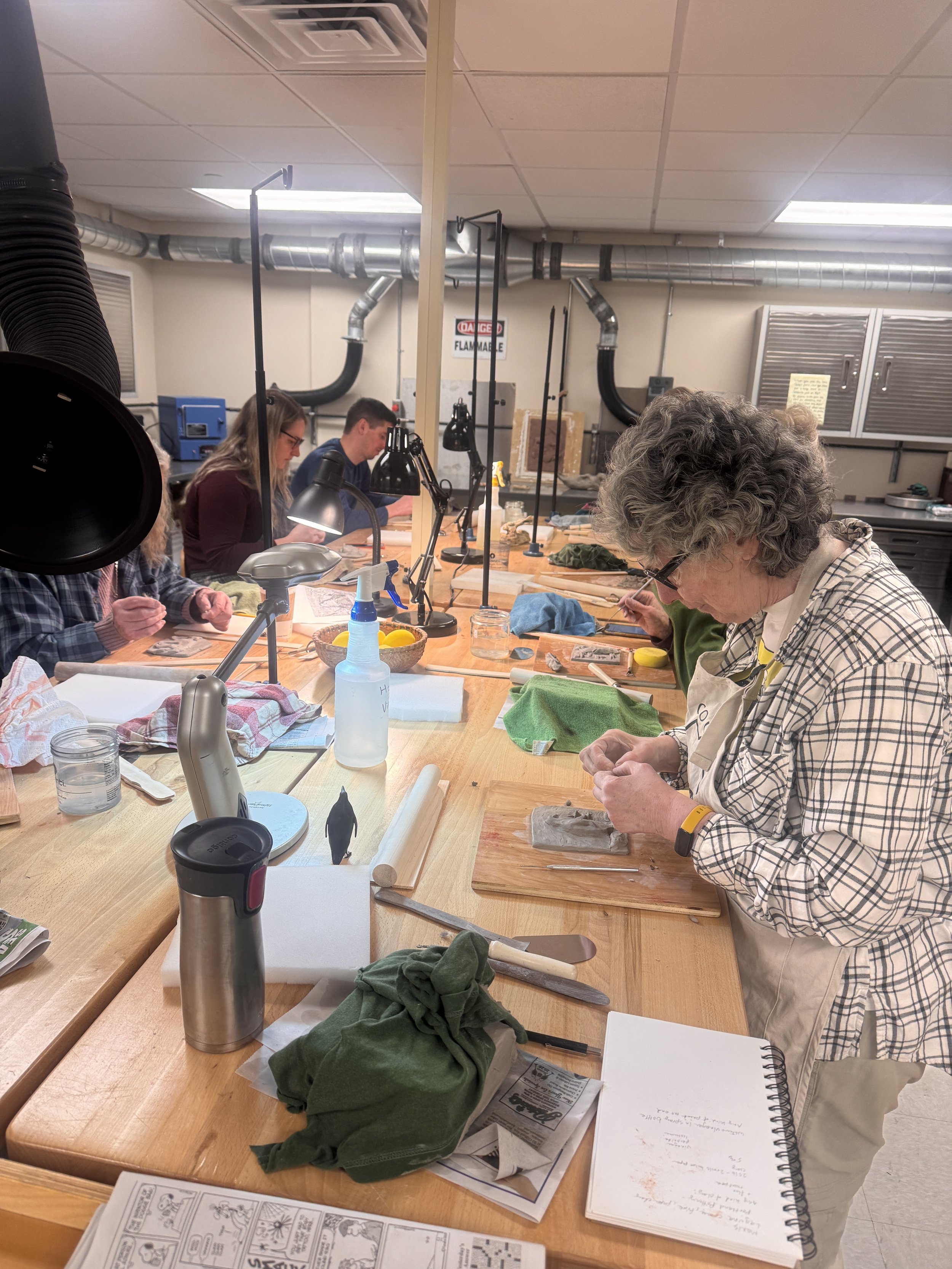 People working at a long wooden table in a well-lit workshop or studio, with various tools, papers, and supplies scattered on the surface. There are large duct pipes, cabinets, and equipment in the background.