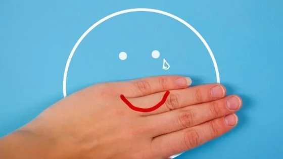 smiley painted hand over sad face indicating masking emotions.