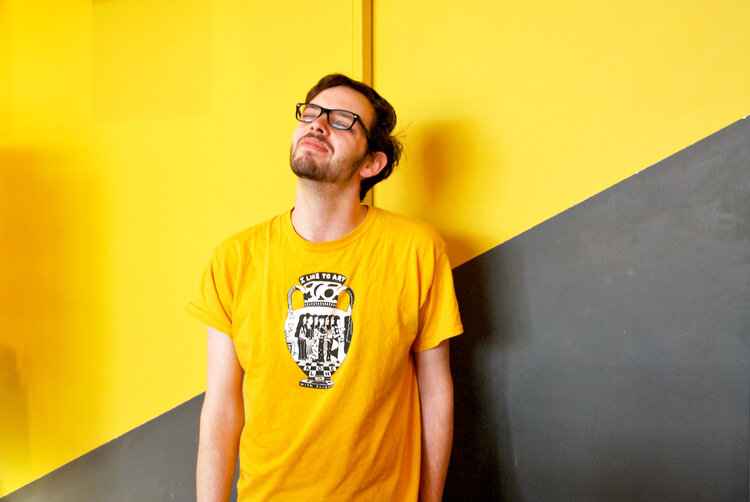 Eli, an employee of Rala, stands smilng in front of a wall in Rala wearing a bright yellow shirt matching the wall behind him.