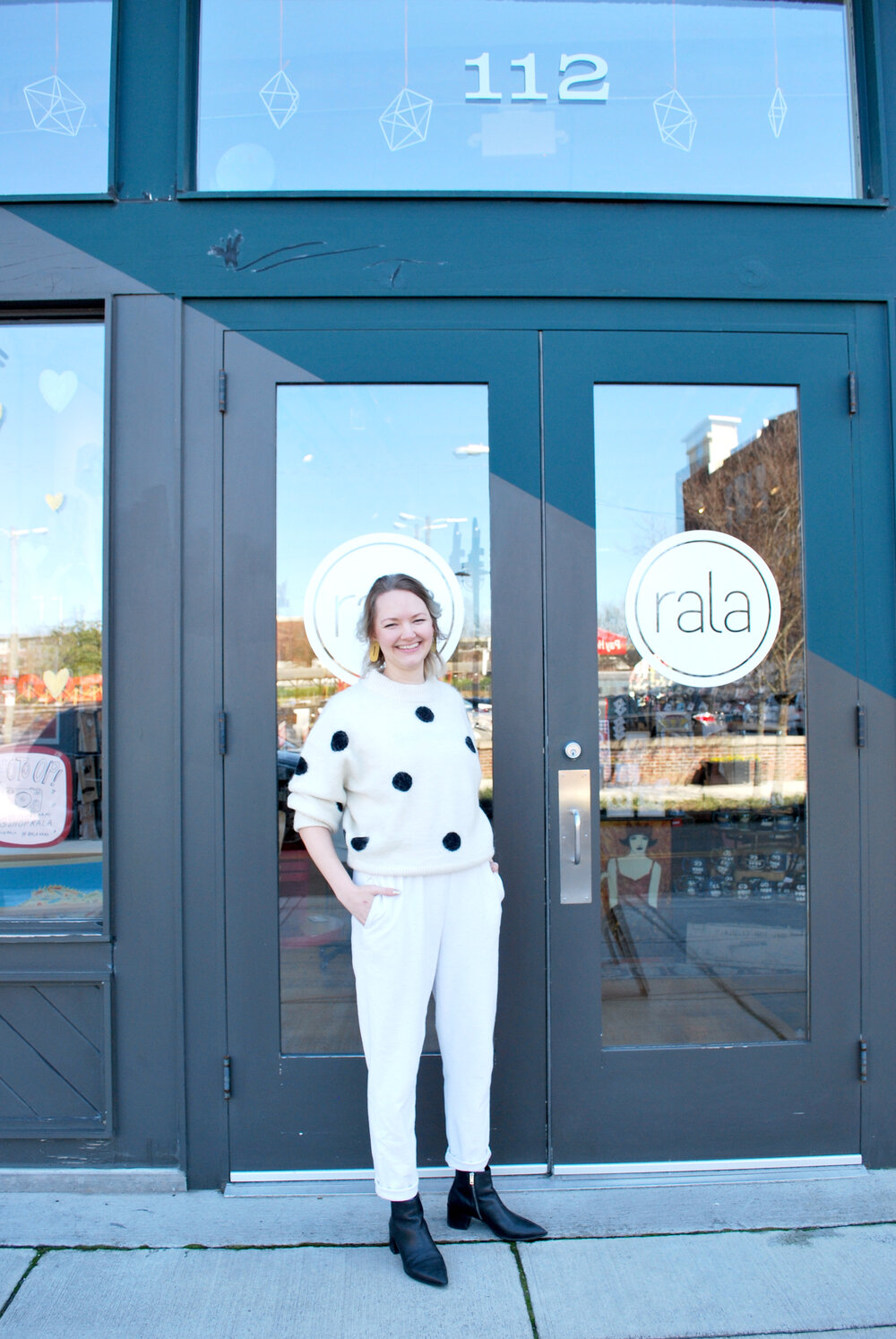 Alaina of Cold Gold standing in front of the Rala doors, wearing a black and white sweater, white pants, and black leather ankle boots.