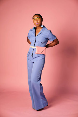 A young woman poses against a pink background in a blue jumpsuit with a pink leather fanny pack at her natural waist.
