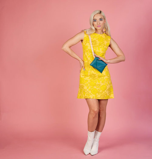 A blonde model wears a yellow sleeveless dress and white boots against a pink background with a leather bely and small turquoise leather bag worn crossbody. 