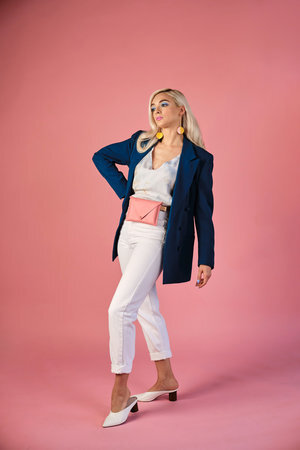 Model poses wearing a Bubblegum pink belt bag with a natural rawhide belt around her waist over white pants and a tie dye tank. 