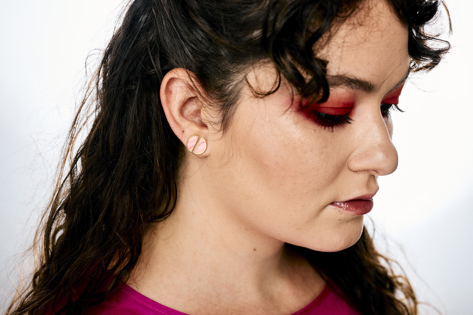 A model with dark curly hair and dramatic red eyeshadow wears the rose quartz half moon geo studs stacked together
