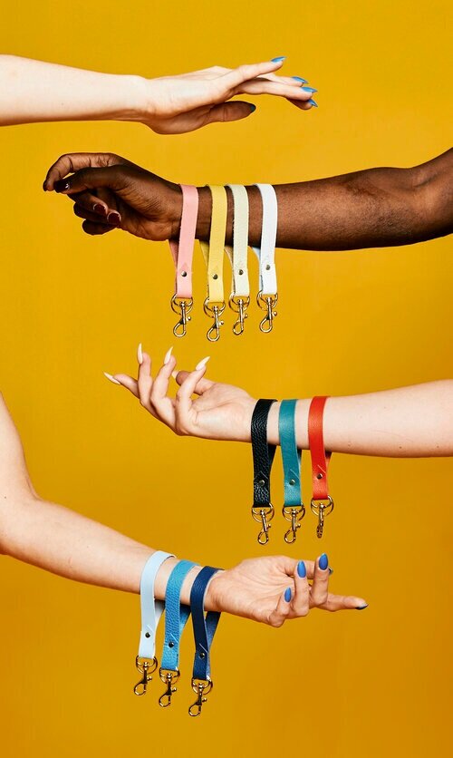 Hand-riveted leather keychains in saturated rainbow colors dangling from arms against a yellow background.