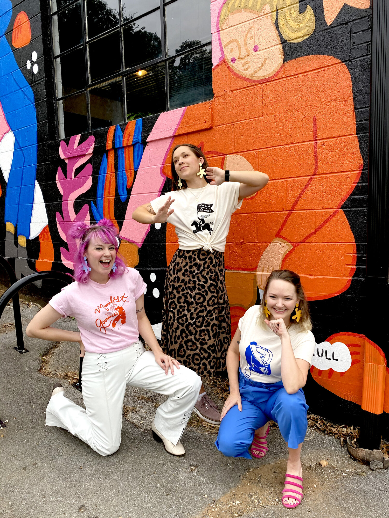 (Left to Right) Shelby of Cold Gold, Paris Woodhull, and Alaina of Cold Gold pose in front of Paris’ mural, all wearing Cold Gold Point Move Earrings and Paris Woodhull t-shirts.