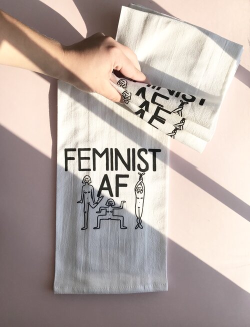 Tea towels designed by Paris Woodhull that say “Feminist AF“ and feature illustrations of 3 nude dancing ladies.