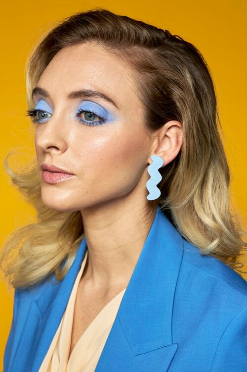 A blonde model wears a perwinkle light trace leather earring with a bright blue suit.