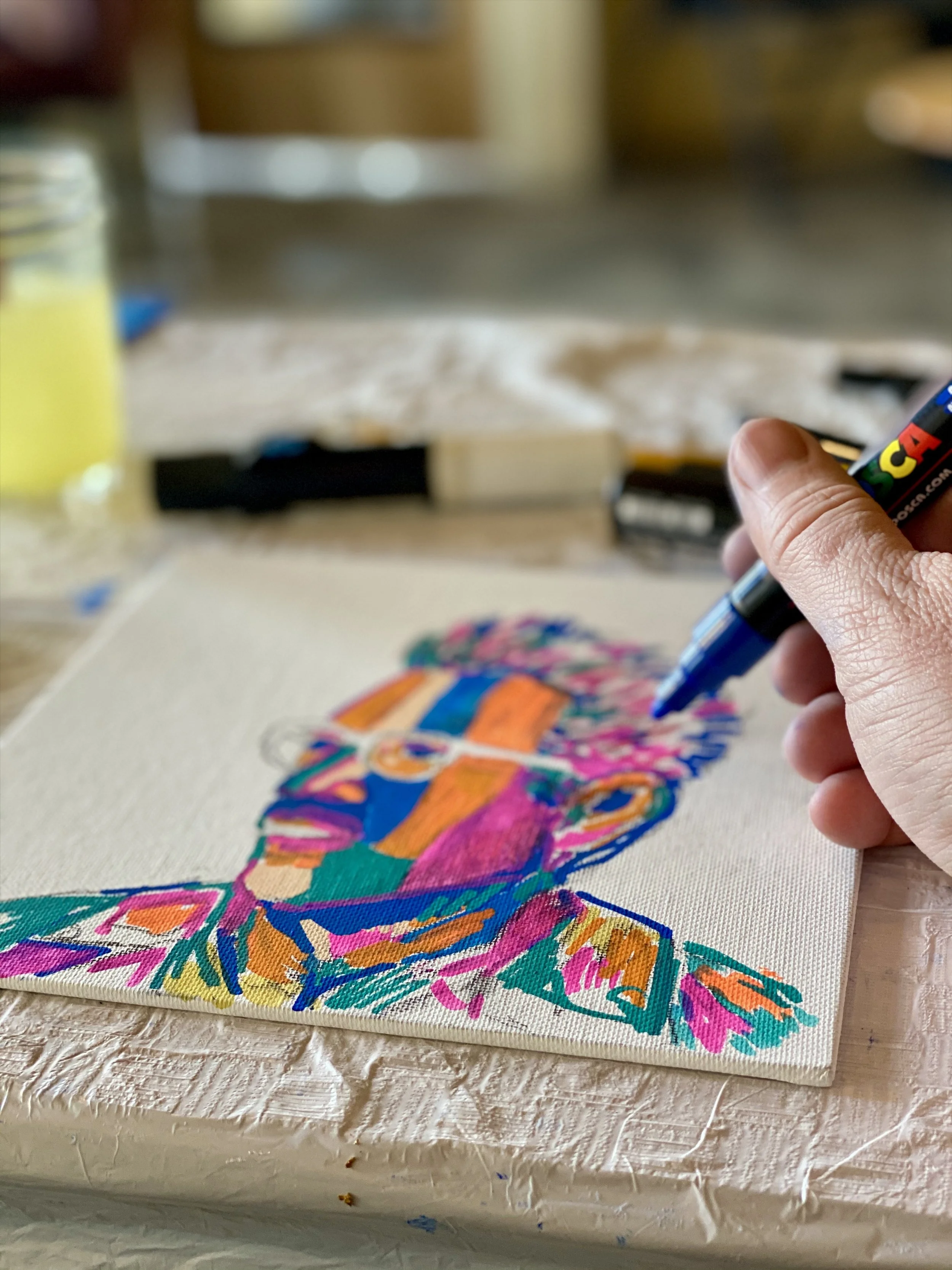 Close-up of a person's hand holding a black marker, drawing on a colorful fabric art piece with bright pink, orange, blue, green, and purple sections, on a white textured surface.